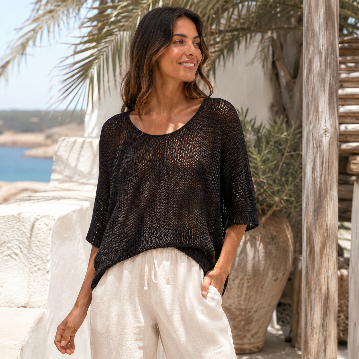 Model trägt schwarzen, luftigen Lochstrickpullover von Marta du Château mit 3/4-Ärmeln kombiniert zu einer weißen Leinenhose in sommerlicher Küstenkulisse.