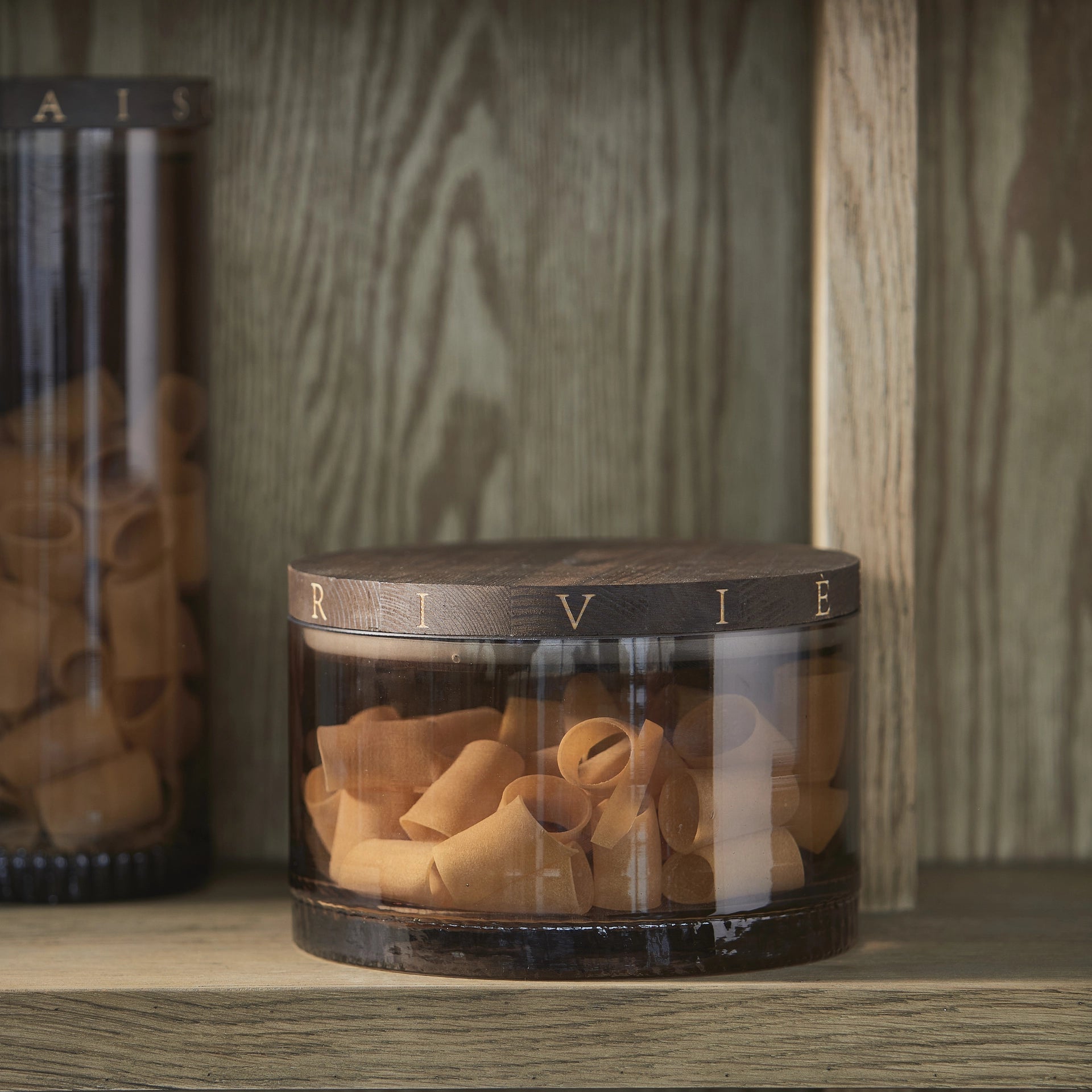 Riviera Maison River Storage Jar aus getöntem Glas mit Holzdeckel, gefüllt mit Pasta auf einer hellen Küchenarbeitsplatte.