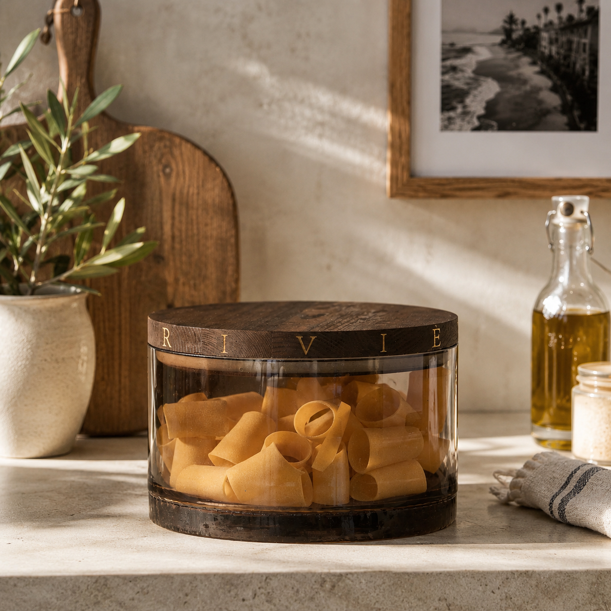 Riviera Maison River Storage Jar aus getöntem Glas mit Holzdeckel, gefüllt mit Pasta auf einer hellen Küchenarbeitsplatte.
