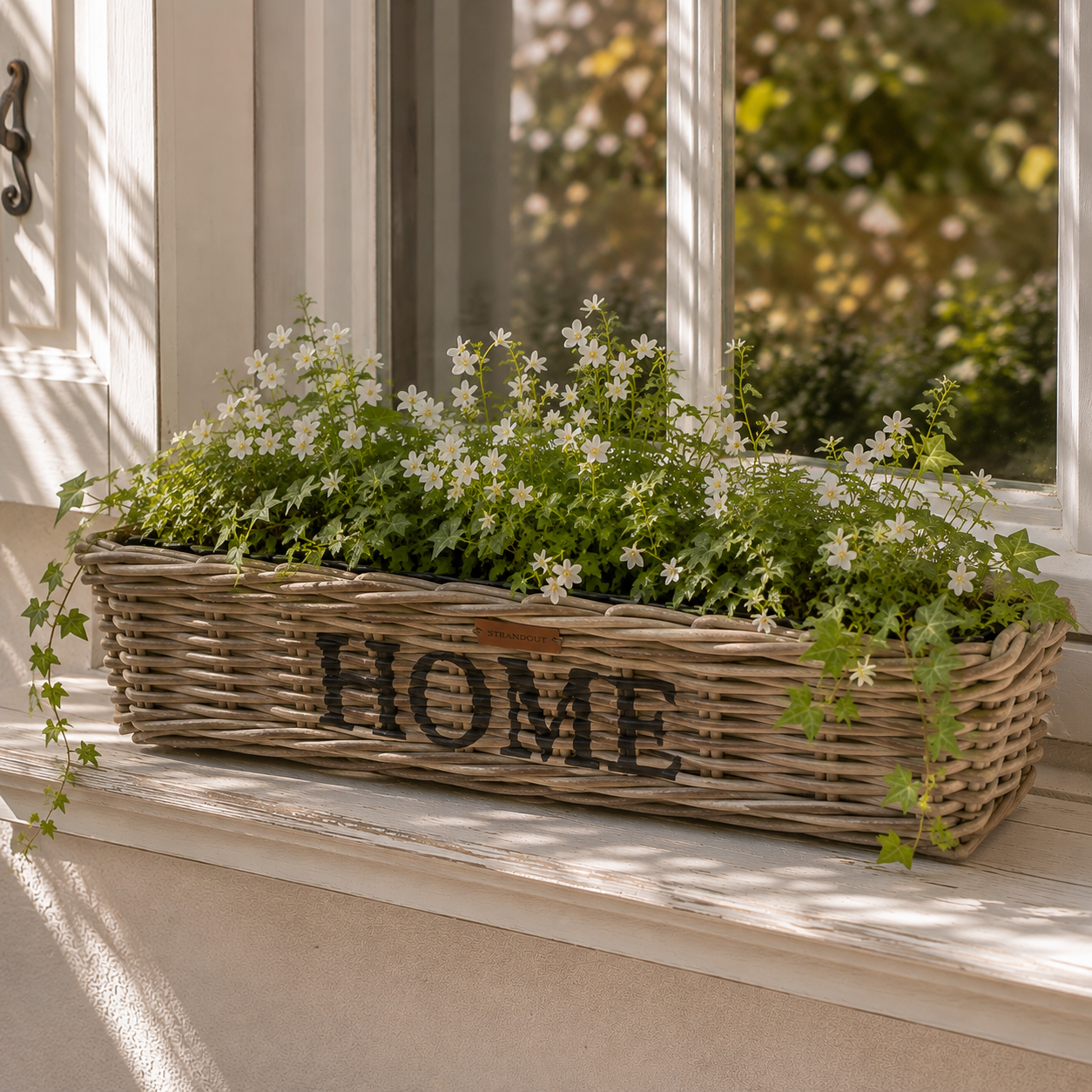 Rattan Planter HOME Größe L mit weißen Blumen und Grünpflanzen auf einer Fensterbank im Landhausstil