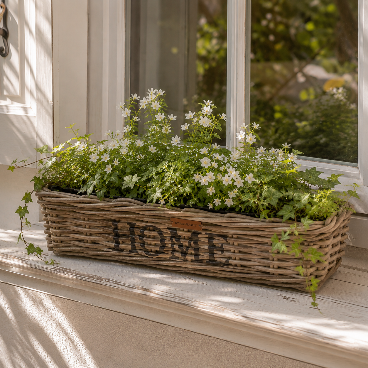 Rattan Planter HOME Größe M mit weißen Blumen und Grünpflanzen auf einer Fensterbank im Landhausstil