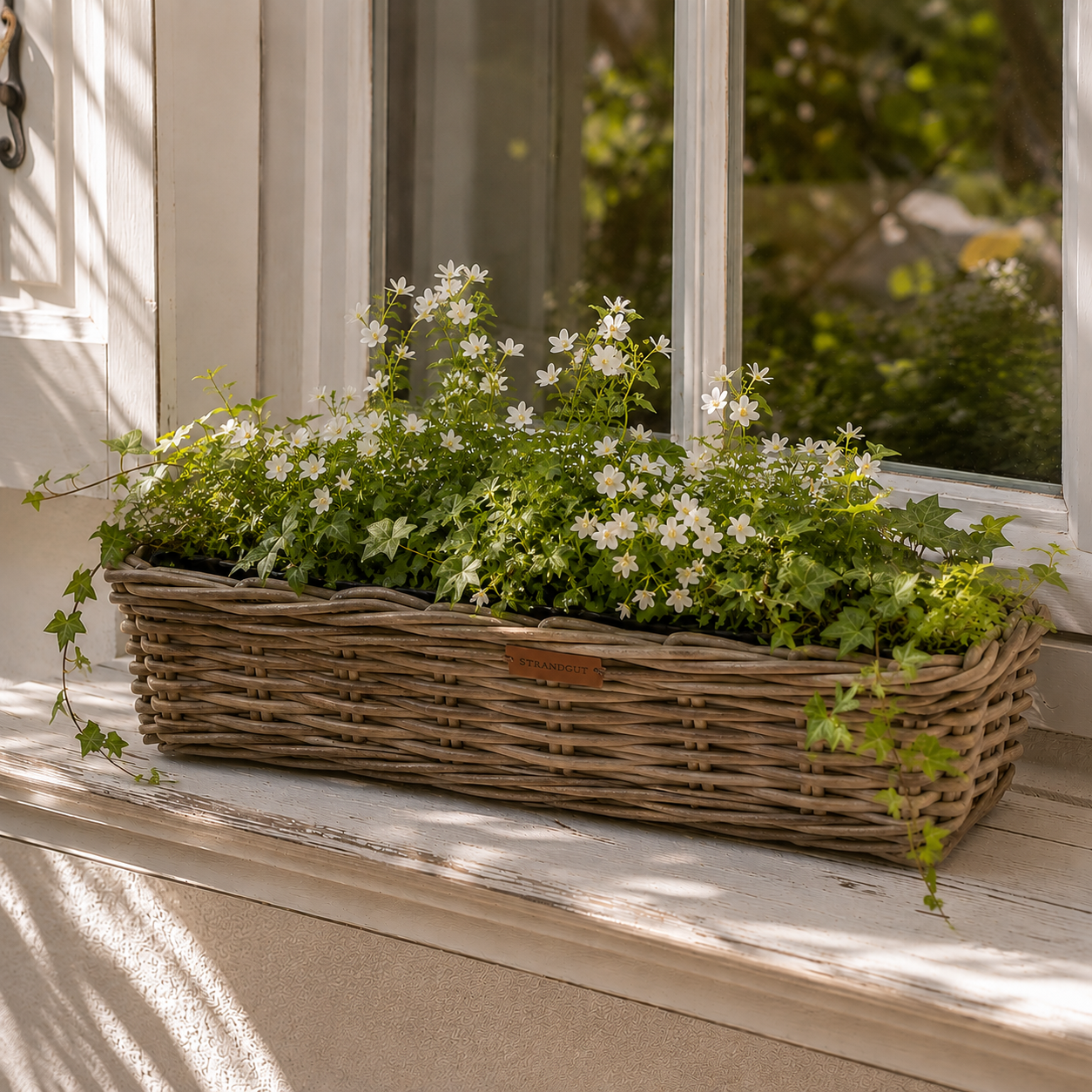 Rattan Planter Rectangular – rechteckiger Pflanzkorb aus natürlichem Rattan mit weißen Blüten und Efeu auf einer hellen Fensterbank