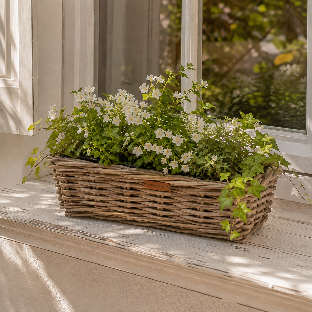 Rattan Planter Rectangular – rechteckiger Pflanzkorb aus natürlichem Rattan mit weißen Blüten und Efeu auf einer hellen Fensterbank