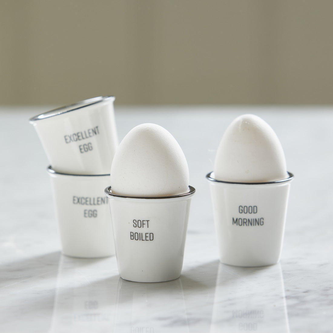 Riviera Maison RM Egg Holder 4 Pieces aus weißem Porzellan mit vier unterschiedlichen Schriftzügen.